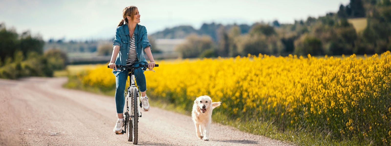 Frau fährt mit ihrem Fahrrad auf einem Schotterweg zwischen Feldern hindurch und neben ihr läuft ein Hund 